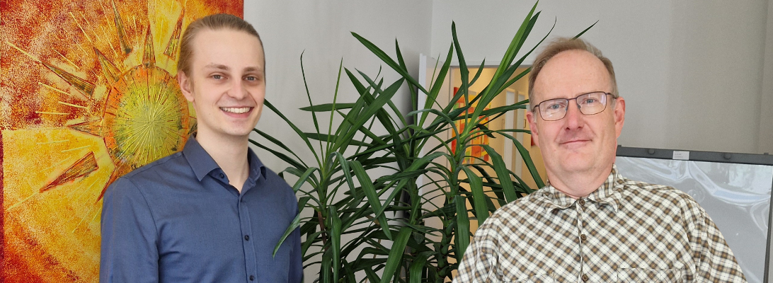 Tim, der bei AKQUINET sein duales Studium absolviert hat, steht neben Holger im Büro. Im Hintergrund ein Bild vom Atelier Lichtzeichen und eine Pflanze.